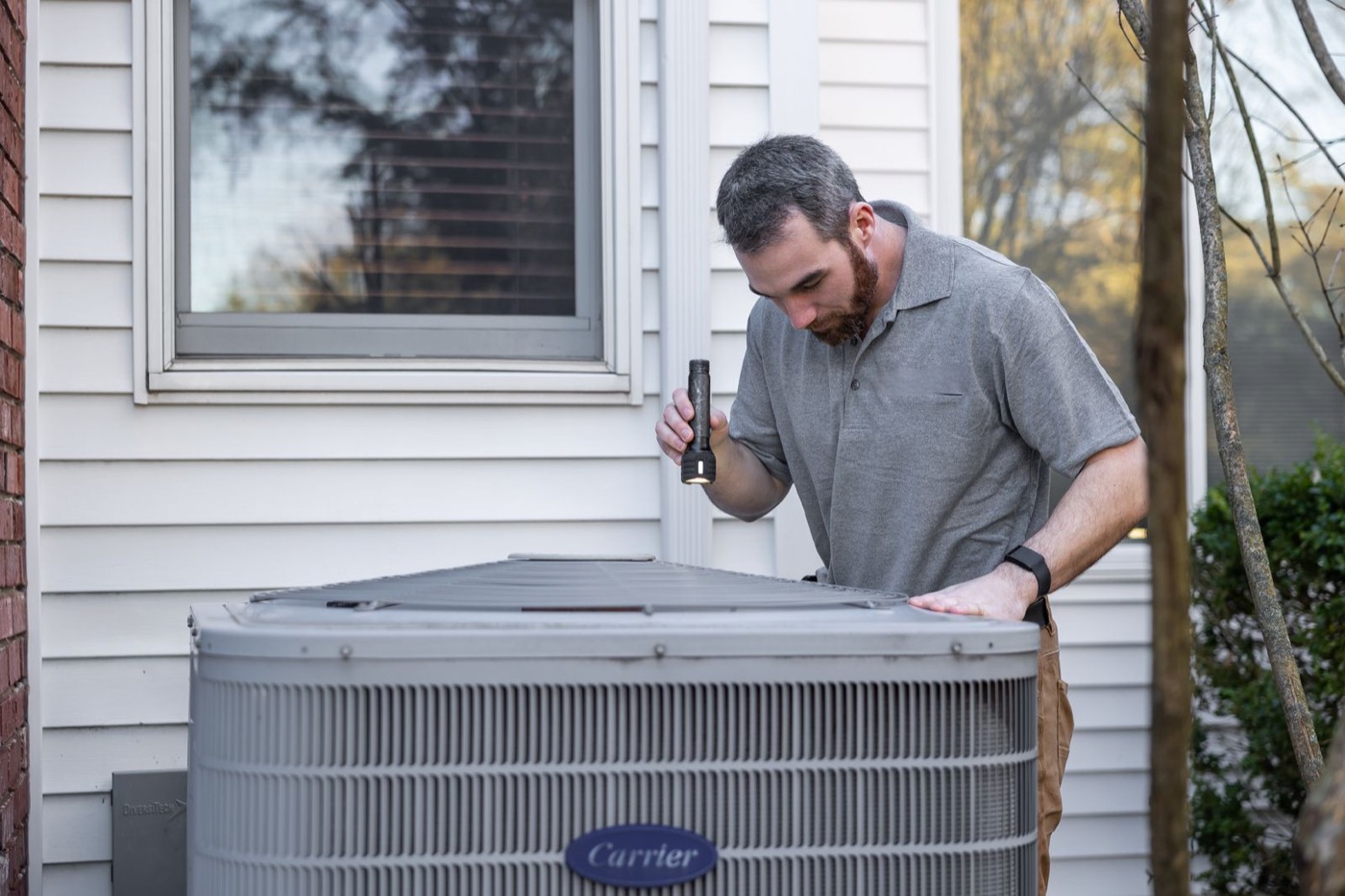 A technician with a flashlight inspecting the top of an outdoor air conditioning unit beside a house